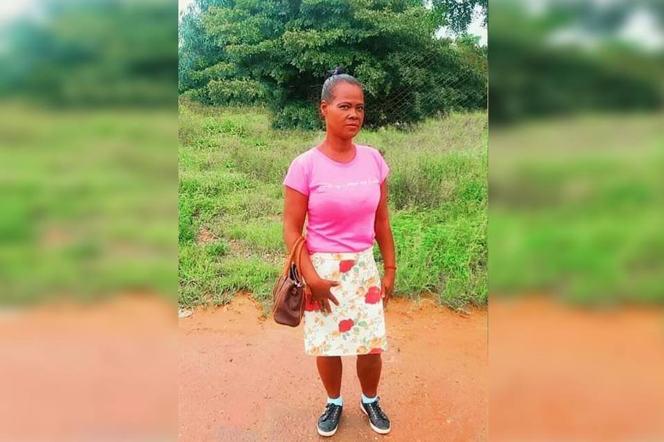 Foto colorida de mulher negra. Ela está em um caminho de terra de chão, veste uma saia com flores e e com uma blusa rosa